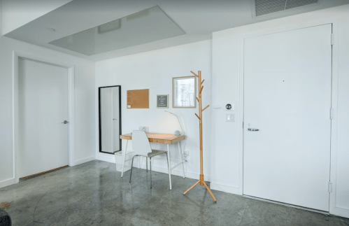 A minimalist room featuring a desk, chair, mirror, coat rack, and a door, with concrete flooring and white walls.
