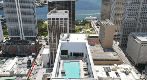 Aerial view of a cityscape featuring buildings, a rooftop pool, and a waterfront in the background.