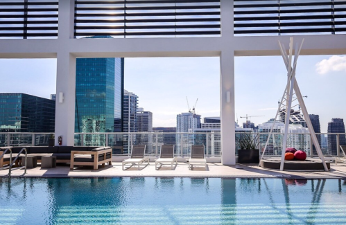 Rooftop pool with lounge chairs and city skyline in the background, featuring modern buildings and clear blue skies.