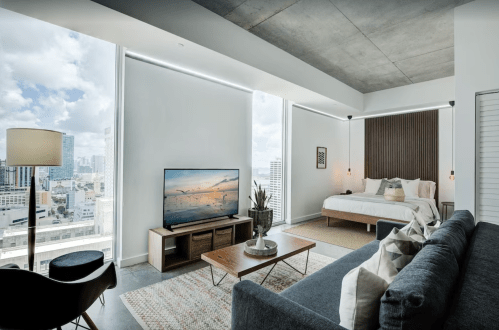 Modern apartment interior with a bed, sofa, TV, and large windows showcasing a city view. Bright and stylish design.