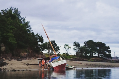 A small boat is stranded on a sandy shore, with people gathered around it and trees in the background.