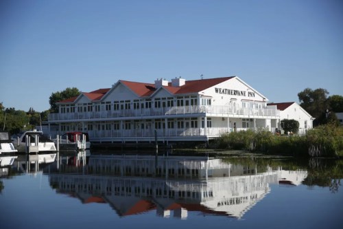 A white inn with a red roof reflected in calm water, surrounded by greenery under a clear blue sky.