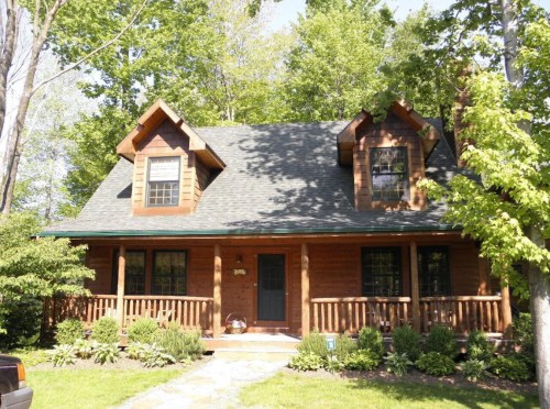 A charming wooden cabin surrounded by trees, featuring a front porch and a sloped roof under a clear blue sky.