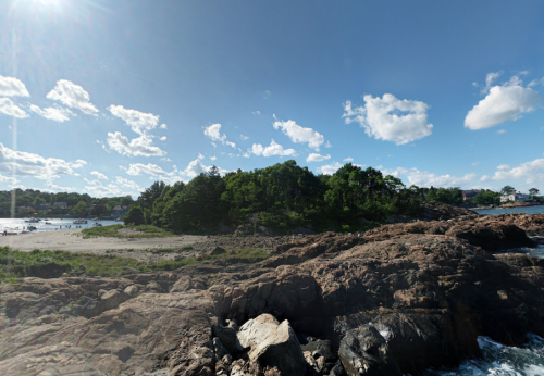A rocky shoreline with lush greenery and a clear blue sky, featuring scattered clouds and a calm sea in the background.