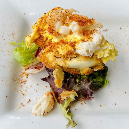 A serving of baked seafood casserole on a bed of mixed greens, topped with seasoning and garnished with crab meat.