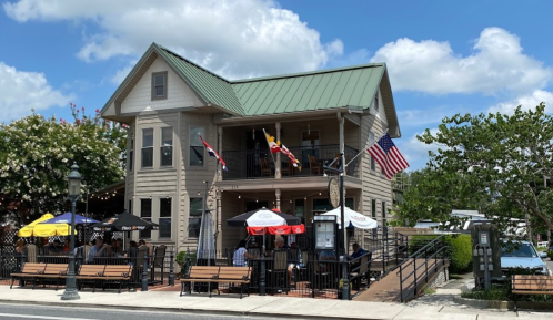 A two-story restaurant with a green roof, outdoor seating, and flags, set against a blue sky with fluffy clouds.