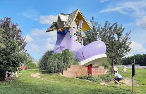 A large purple shoe-shaped structure with a roof, surrounded by greenery and playful decorations, under a blue sky.