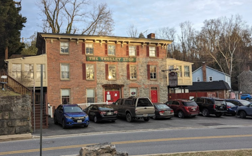 A brick building labeled "The Trolley Stop" with parked cars in front, set against a wooded backdrop.