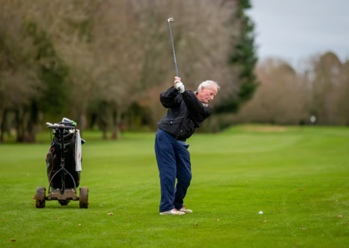 A golfer swings a club on a lush green course, with a golf bag on a cart nearby.