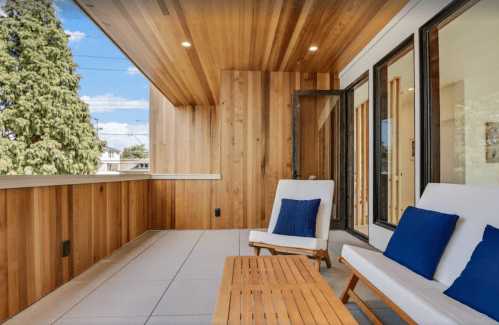 A modern balcony with wooden walls, two white chairs with blue cushions, and a wooden table under a clear sky.