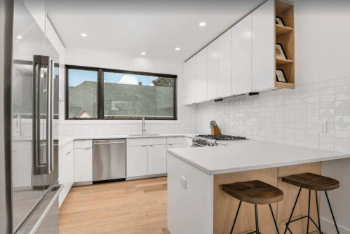 Modern kitchen with white cabinets, stainless steel appliances, and a large window overlooking a roof. Two wooden stools at the island.