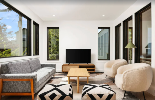 Modern living room with large windows, a gray sofa, two armchairs, a coffee table, and a TV on a wooden stand.