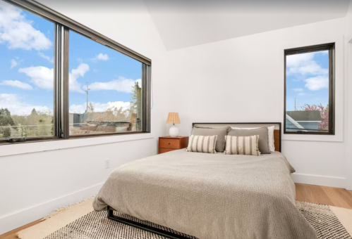 Bright bedroom with a bed, decorative pillows, a nightstand, and large windows showcasing a blue sky.