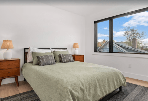 Cozy bedroom with a green bedspread, two lamps, and a large window showing a view of rooftops and blue sky.