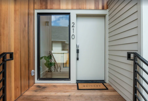 Front entrance of a modern home featuring a white door, large window, wooden accents, and a welcome mat.