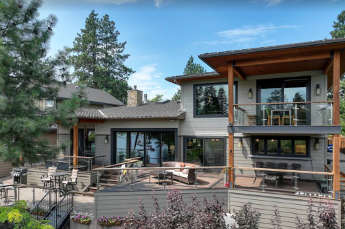 Modern lakeside home with large windows, a spacious deck, and surrounding trees. Outdoor seating area visible.