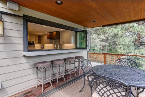 A cozy outdoor dining area with a view of a kitchen window and bar stools, surrounded by trees.