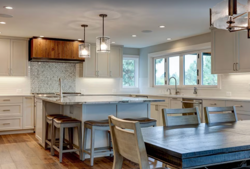 Modern kitchen with an island, wooden accents, pendant lights, and a dining table, featuring light cabinetry and large windows.