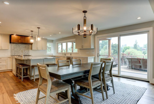 A modern kitchen and dining area with a wooden table, chairs, and large windows overlooking greenery.
