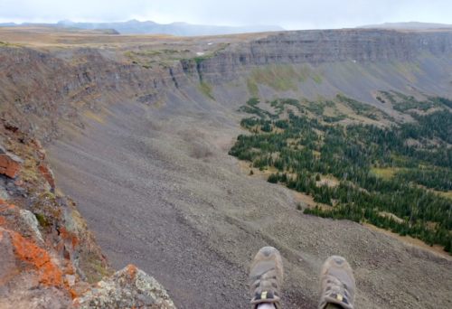 A pair of feet in hiking shoes dangling over a rocky cliff, overlooking a vast, green valley below.