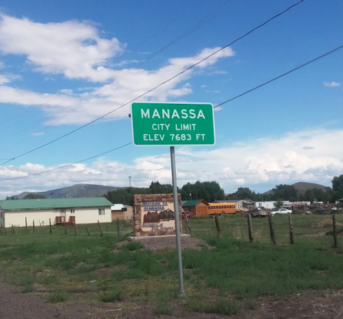 Sign marking the city limit of Manassa, elevation 7,683 feet, with a blue sky and mountains in the background.