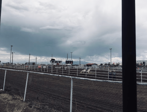 A rodeo arena under a cloudy sky, with spectators and a fenced dirt area in the foreground.