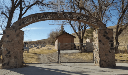 Archway entrance to a cemetery with a cross, surrounded by trees and a fenced path leading to a building.