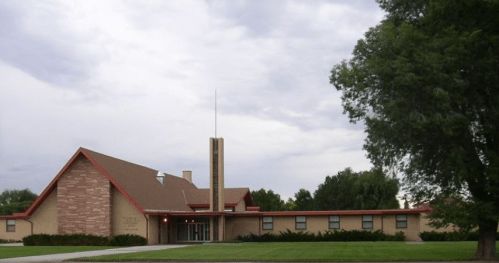 A modern brick building with a sloped roof, tall chimney, and grassy area, set against a cloudy sky.