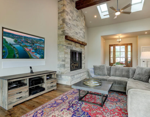 Cozy living room with a stone fireplace, large TV, sectional sofa, and a decorative rug. Bright natural light from windows.