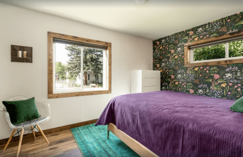 Cozy bedroom with a purple bedspread, floral wallpaper, and a window overlooking greenery.