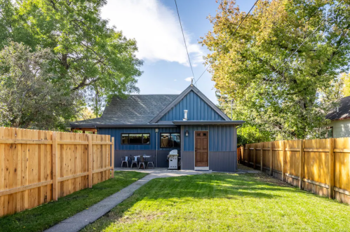 A blue house with a wooden fence, surrounded by trees, featuring a small patio and grill in the backyard.