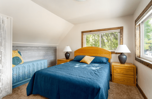 Cozy bedroom with a blue bedspread, wooden furniture, and large windows letting in natural light.