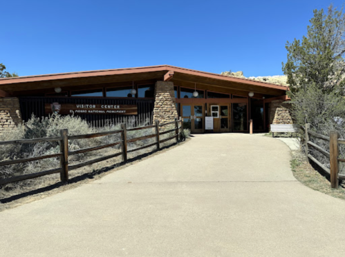 Visitor center building with stone accents, surrounded by greenery and a pathway leading to the entrance. Clear blue sky.
