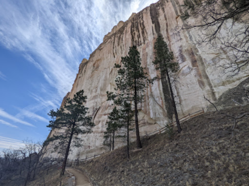 A path lined with trees leads to a towering white cliff under a blue sky with wispy clouds.
