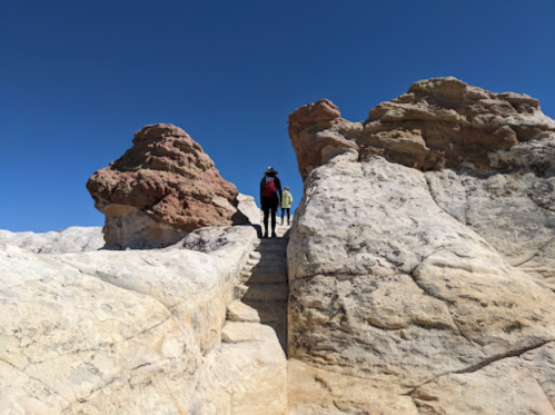 Two hikers ascend rocky terrain under a clear blue sky, surrounded by unique rock formations.