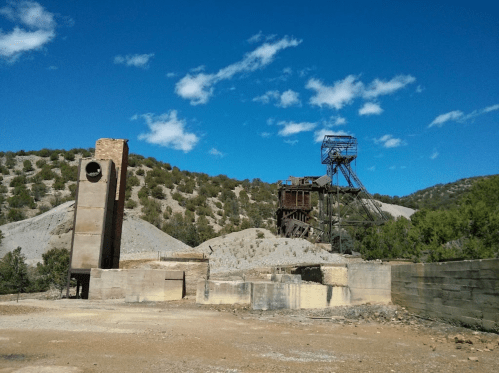 Abandoned mining site with a tall chimney and machinery against a blue sky and green hills.
