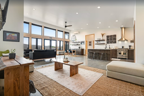 Modern living room with large windows, minimalist furniture, and an open kitchen, featuring a wooden coffee table and area rugs.