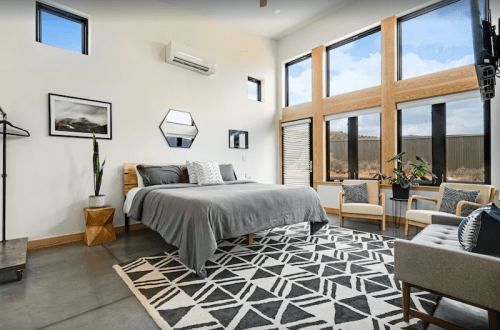 Modern bedroom with large windows, a gray bed, geometric rug, and stylish seating. Bright and airy atmosphere.