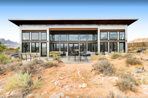 Modern house with large windows, surrounded by desert landscape and rocky formations under a clear blue sky.