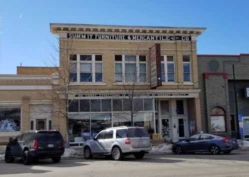 Historic building with "Summit Furniture & Mercantile Co" sign, featuring large windows and parked cars in front.