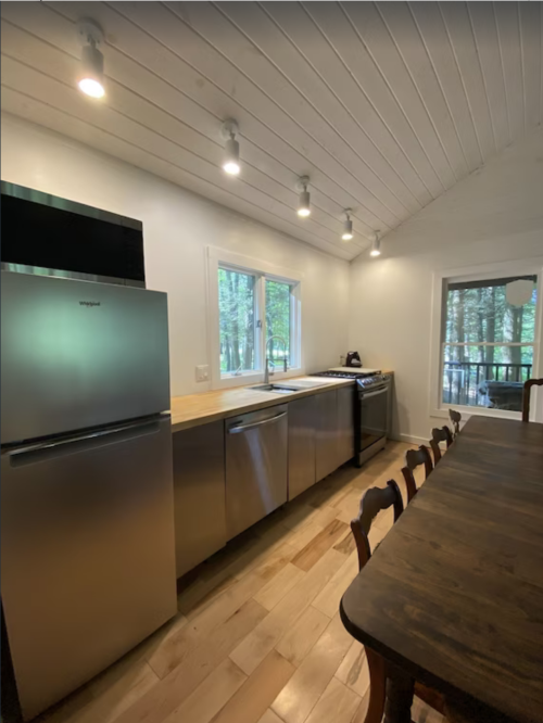 Modern kitchen with stainless steel appliances, wooden countertops, and a dining table, surrounded by large windows.