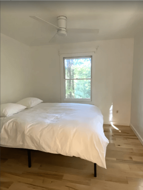 A bright, minimalist bedroom featuring a bed with white bedding and a window overlooking greenery.