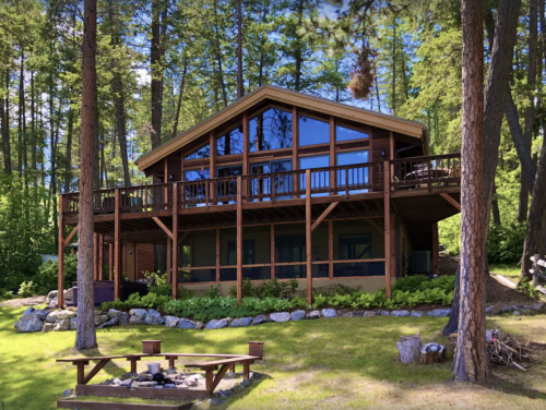 A modern cabin surrounded by trees, featuring a large deck and a fire pit in the foreground.
