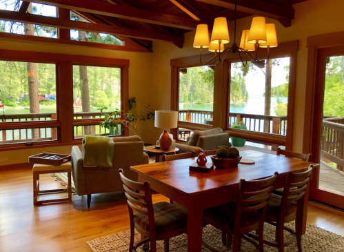 Cozy living space with large windows, wooden furniture, and a view of a lake surrounded by trees.
