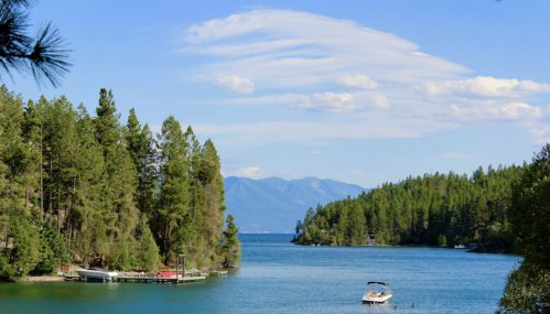 A serene lake surrounded by lush green trees and mountains, with a boat on the water under a blue sky.