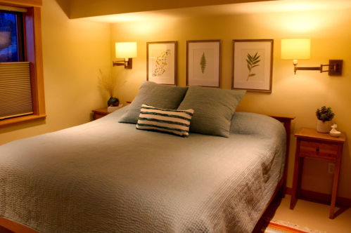 Cozy bedroom with a large bed, decorative pillows, and framed botanical art on the walls, softly lit by wall sconces.