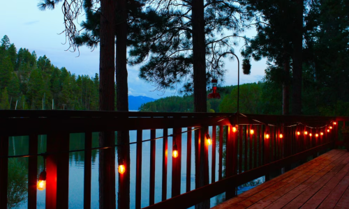 A serene lakeside view at dusk, with string lights illuminating a wooden deck surrounded by tall trees.