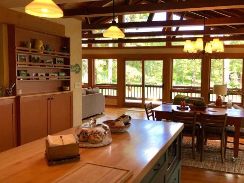 Bright, open kitchen with wooden accents, a dining area, and large windows overlooking a forested view.