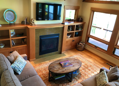 Cozy living room with a fireplace, TV, wooden shelves, and large windows overlooking a deck. Warm, inviting decor.