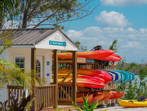 A water sports rental hut with colorful kayaks stacked outside, surrounded by greenery and a blue sky.